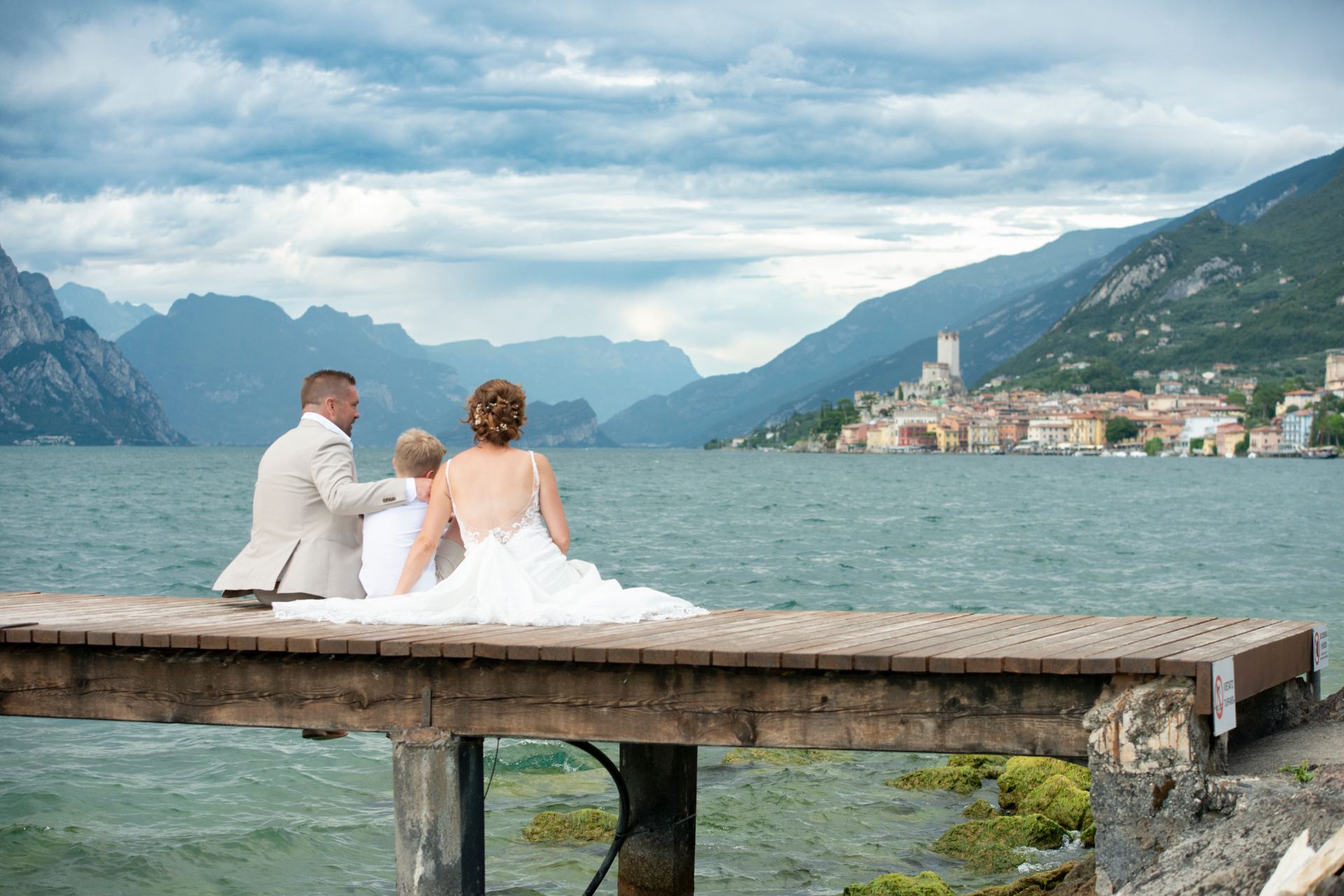 Couple sitting on a wooden jetty looking across Lake Garda toward Malcesine